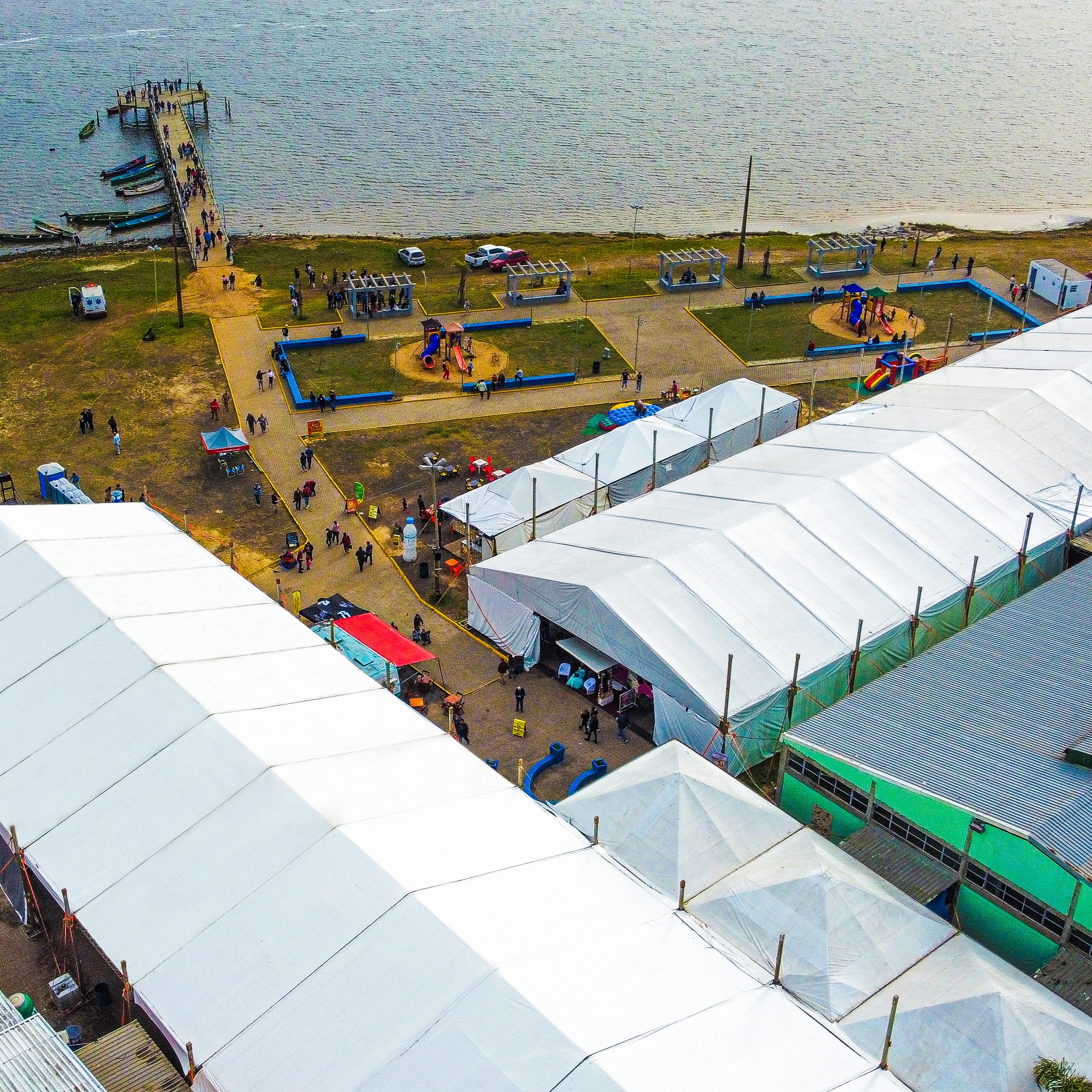 Vista aérea de feira de eventos com pavilhões brancos montados lado a lado, público circulando entre estandes, orla de lago ao fundo com deck de madeira e barcos, paleta natural verde e branco, luz difusa de fim de tarde, ângulo de drone, fotografia realista